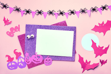 Creative Halloween flat lay background on pink paper with copy-space. Blank card and paper decorations: bats, and jack lantern pumpkins. Decorative garland with flags and spiders.