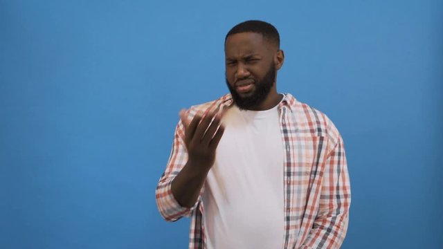 Afro american man isolated against blue background smelling something stinky and disgusting, intolerable smell, holding breath with fingers on nose. Bad smells concept. 4K