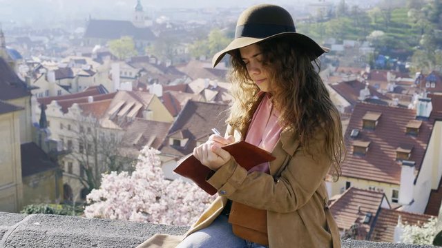 Woman in brown hat writing in the diary on view point with amazing view. Portrait concentrated girl taking notes and drawing in paper notepad while sitting on parapet above beautiful cityscape 