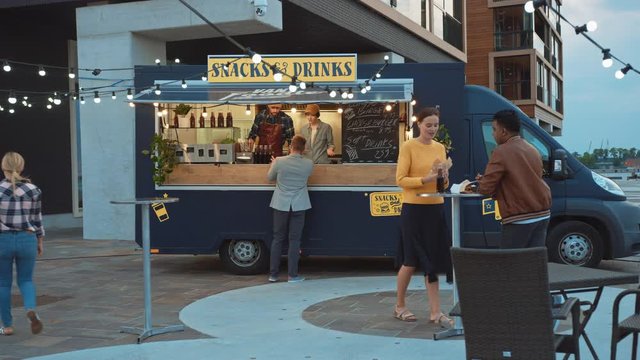 Food Truck Employee Hands Out Freshly Made Beef Burgers, Fries and Cold Drinks to Happy Young Hipster Customers. Commercial Truck Selling Street Food in a Modern Cool Neighbourhood.