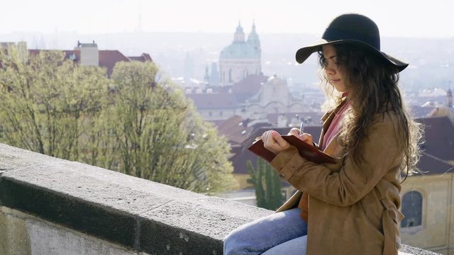 Woman in brown hat writing in the diary on view point with amazing view. Portrait charming girl taking notes and drawing in paper notepad while sitting on parapet above beautiful cityscape 