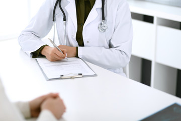 Woman doctor and patient sitting and talking at medical examination at hospital office, close-up. Therapist filling up medication history records. Medicine and healthcare concept