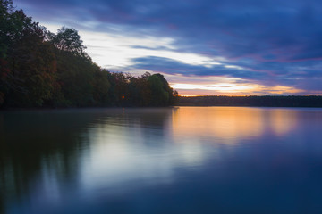Obraz premium Landscape; lake and trees before sunrise, long exposure blurred objects, dark clouds and sky, autumn trees and orange sun are reflected in the water