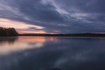 Naklejka na ściany i meble Landscape; lake and trees before sunrise, long exposure blurred objects, dark clouds and sky, autumn trees and orange sun are reflected in the water