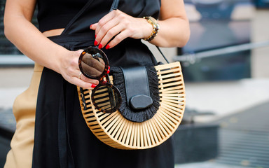 Fashionable bag close-up in female hands.Girl walks in the city outdoors. Stylish modern and feminine image, style. Woman in a dress and beige trousers, holds a straw or wooden bag. Sunglasses.