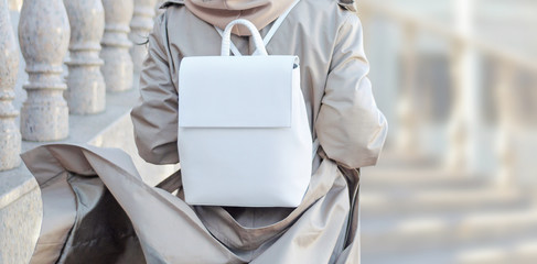 Fashionable bag close-up in female hands.Girl walks in the city outdoors. Stylish modern and feminine image, style. Woman in a raincoat, coat and with a white backpack on his shoulder. girl backs.