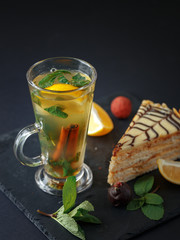 Warming herbal tea with lemon and cinnamon on a black slate plate with a piece of cake, slices of lemon and mint.
