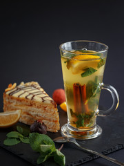 Warming herbal tea with lemon and cinnamon on a black slate plate with a piece of cake, slices of lemon and mint.