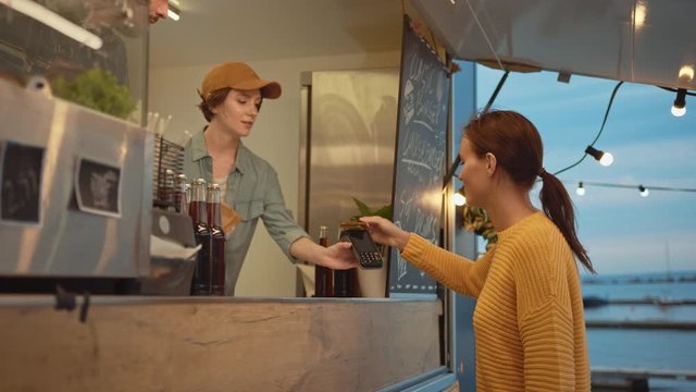 Food Truck Employee Hands Out a Freshly Made Burger to a Happy Young Female. Young Lady is Paying for Food with Contactless Credit Card. Street Food Truck Selling Burgers in a Modern Hip Neighbourhood
