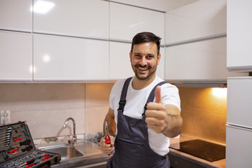 Portrait of a handyman in uniform standing in the renovated kitchen at home showing thumb up. Young worker with tool bag standing near oven in kitchen