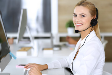 Fototapeta na wymiar Blonde business woman using headset for communication and consulting people at customer service office. Call center. Group of operators at work at the background