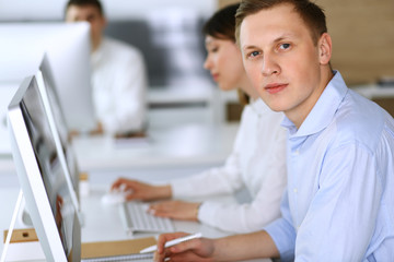 Cheerful smiling businessman headshot at work in modern office. Unknown casual dressed entrepreneur using pc computer while sitting with diverse colleagues at the background. Multi ethnic working