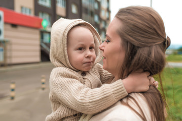 Young mother woman holding a little boy 3-5 years old in her arms, a beige sweater warm clothes. Emotions of joy of love and fun play. Cold weather outside, in hood.