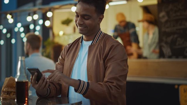 Handsome Young Indian Man is Using a Smartphone while Sitting at a Table in a Outdoors Street Food Cafe