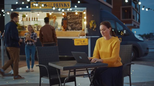 Beautiful Brunette Young Woman is Working on Laptop while Sitting at a Table in an Outdoors Street Food Cafe. She's Browsing Internet or Social Media, Posting a Status Update. Waiting for Her Order.