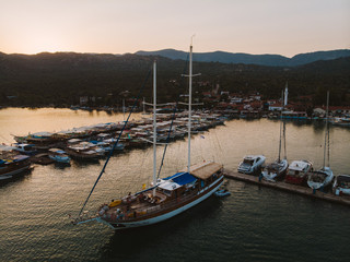 Luxury yachts docked in a remote island bay off the coast of turkey. Sun is setting over the mountains behind the town. Shot aerially from a drone. 