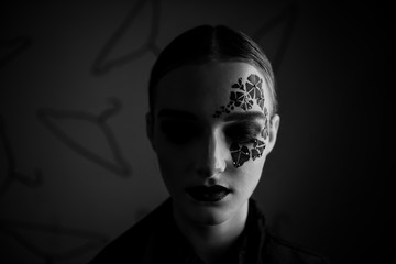 Close-up portrait of a girl with clean skin, bright professional make-up, purple lipstick, face with black ivy tattoo around her eyes with rhinestones. Gray background, pattern of hangers. Fashion