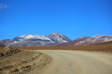 Wide dusty road crossing the desert in the Bolivia highlands. Mountains with snow in the peaks and clear blue sky