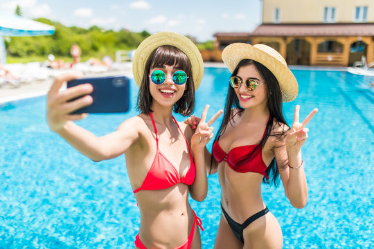Two attractive brunette women wearing bikini posing near the swimming pool, making selfie photo on the phone for social media