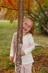 Adorable baby girl playing in a sunny park under a tree with yellow leaves, hiding behind tree