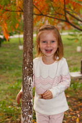 Adorable baby girl playing in a sunny park under a tree with yellow leaves, hiding behind tree