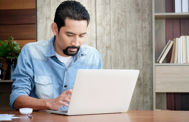 Asian handsome bearded guy sitting at the working desk in his house using a laptop for work. Online business entrepreneur checking the order list on the internet platform.