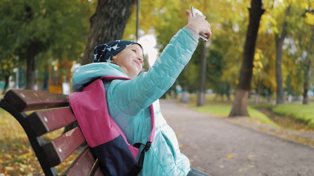 A beautiful teen girl sits on a bench in a park during golden autumn time and takes selfie photos with her mobile phone. Concept of online addiction and social networks.