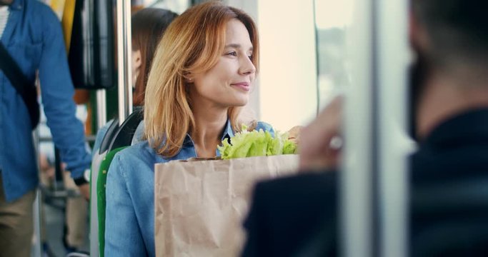 Portrait of the pretty blond Caucasian woman with a big packet of vegetables and other food going home in the tram from the supermarket or market, looking at the window and smiling.