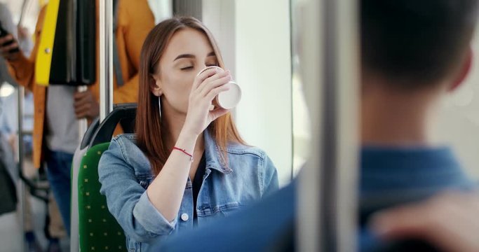 Young Caucasian beautiful girl sitting in the tram while going somewhere, holding a cup to-go and sipping hot coffee while smiling and flirting to the guy in front of her.