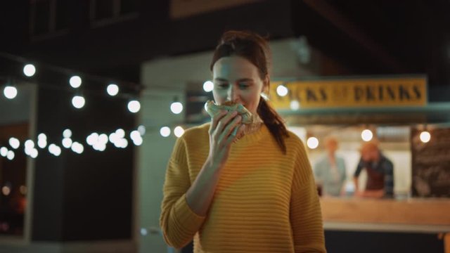 Beautiful Young Female Walks Away from a Food Truck with a Delicious Fresh Beef Burger. She's Happy with Hew Food and Takes a Biet. Commercial Kiosk is Selling Street Food on a Modern Cool Street.