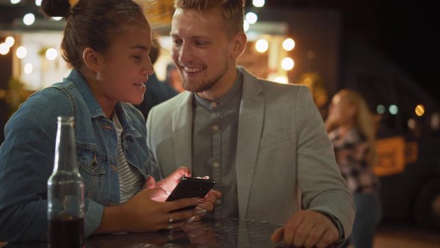 Beautiful Young Couple is Using a Smartphone while Sitting at a Table in an Outdoors Street Food Cafe. They're Browsing Internet or Social Media, Watching Videos. They are Happy and Smile.