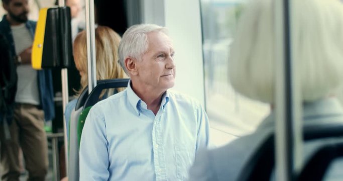 Portrait shot of the handsome gray-haired Caucasian old man going home in the tram and looking at the window, then smiling and talking to the senior woman who sitting in front of him.