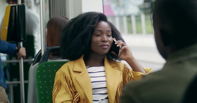 Beautiful young African American girl with curly hair sitting in the tram and talking cheerfully on the mobile phone. Rear of the guy.