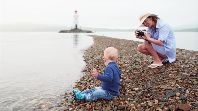 Young mother is taking picture of her little son who is sitting on pebble and throwing stones in water. Tokarevsky lighthouse is on the background. Morning. Vladivostok, Russia