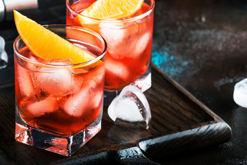 Negroni alcoholic cocktail with dry gin, red vermouth and red bitter, orange slice and ice cubes. Brown bar counter background, bar tools, night mood, copy space, selective focus