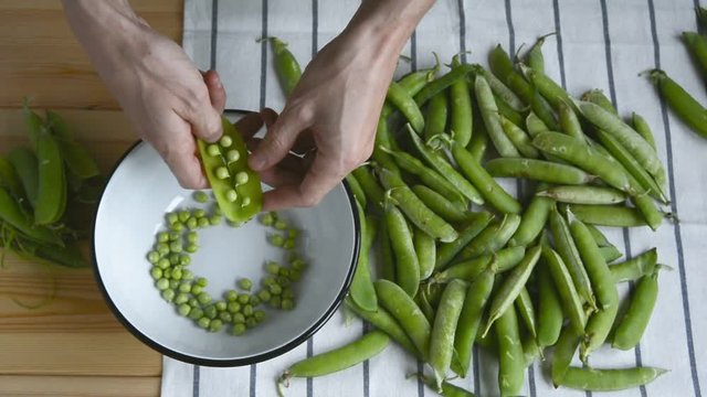 mans hand taking fresh ripe green pea bean from pile on striped napkin, shelling, putting peas into metal bowl pod husk on wood table, top view of closeup full hd stock video footage in real time