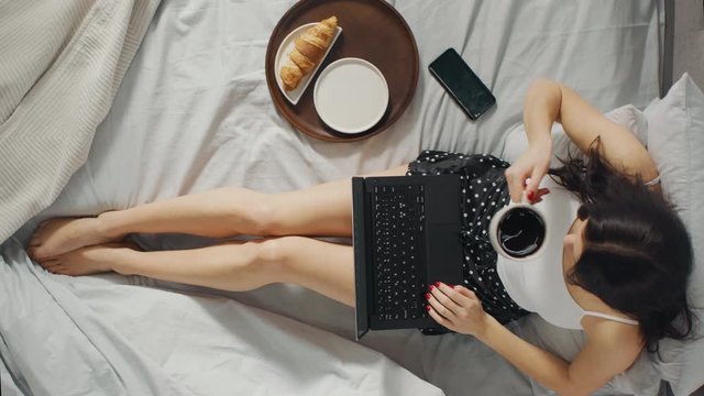 Young Millennial Girl Sitting on a Bed in the Morning, Uses Laptop Computer and Eats Croissants, Drinks Coffee for Breakfast