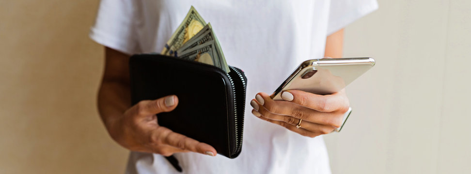 Wallet With US Dollars And Mobile Phone In The Woman Hands, Cash Pay Or Shopping Concept. Web Banner. Girl Get Out The Money From The Black Leather Purse, Hand Holding One Hundred Dollars In A Purse.