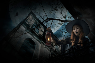 Halloween witch holding ancient lamp standing over grunge castle, dead tree, bird fly, full moon and cloudy spooky sky, Halloween mystery concept