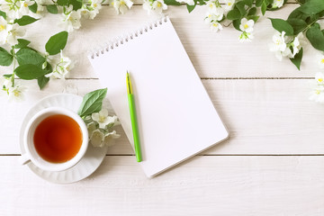 a cup of green tea and Jasmine flowers on a white background top view. morning tea and an empty notebook with a pen on the table. copy space. the cup and flowers lay flat.