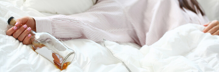 Young woman lying in bed deadly drunken holding near-empty bottle of booze. Female intoxicated with alcohol after tough night party. Alcoholism habitual drunkenness pernicious habit concept