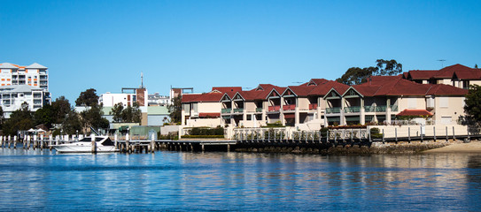 Fototapeta premium Large waterside houses, apartment condominiums in suburban community on riverfront with boat moored at wharf, blue sky in background