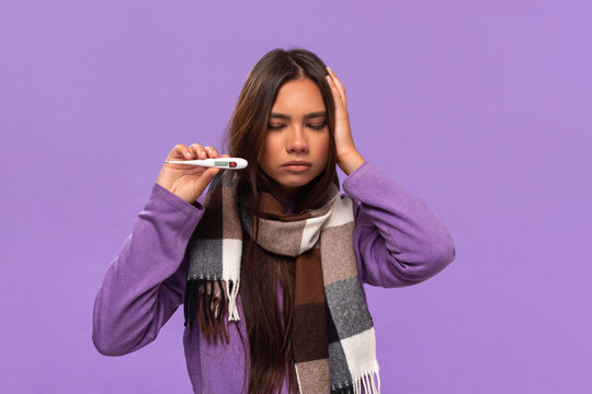 Pretty african-american girl in a sweater and cosy checked scarf measuring thermometer isolated over purple background. Catching flu in winter.