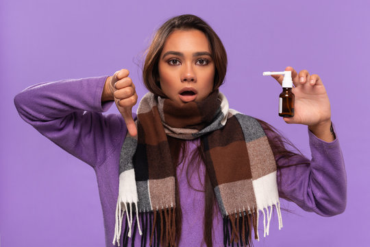 Pretty african-american girl in a sweater and cosy checked scarf got tired of using nose spray beacuase of cold standing isolated over purple background. Catching flu in winter.