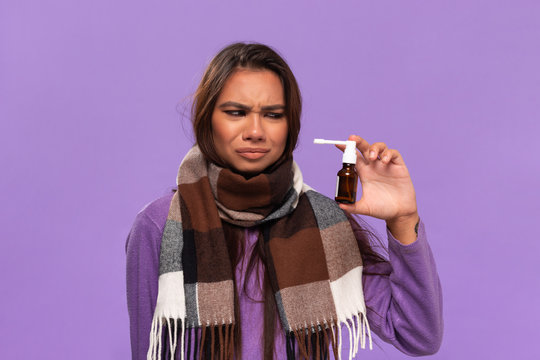 Pretty african-american girl in a sweater and cosy checked scarf got tired of using nose spray beacuase of cold standing isolated over purple background. Catching flu in winter.