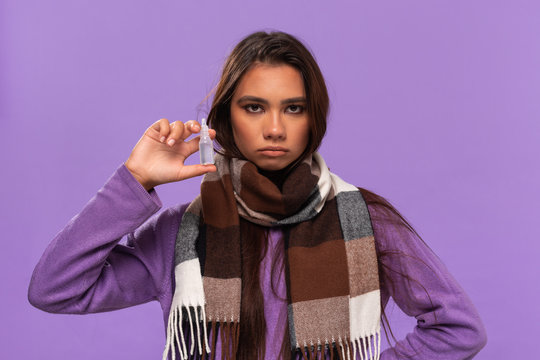Pretty african-american girl in a sweater and cosy checked scarf holding nasal drops to cure common cold standing isolated over purple background. Catching flu in winter.