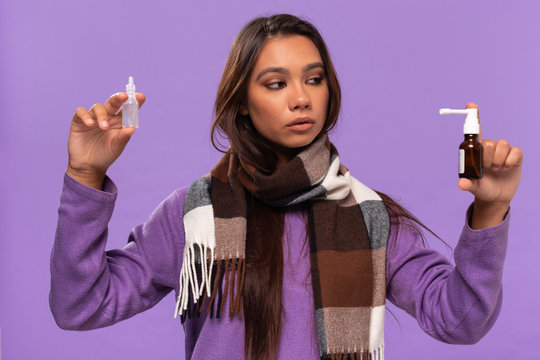 Pretty african-american girl in a sweater and cosy checked scarf holding nasal drops  and spray to cure common cold standing isolated over purple background. Catching flu in winter.