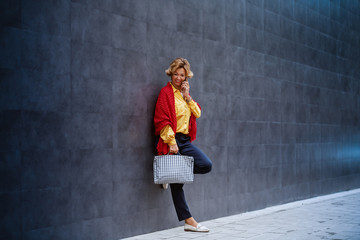 Full length of beautiful caucasian blonde fashionable senior woman with scarf holding bag and talking on the phone while leaning on the wall outside.
