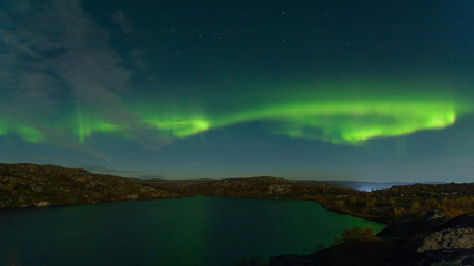 Northern lights, aurora in the sky above the hills and cliffs and reflected in the lake.