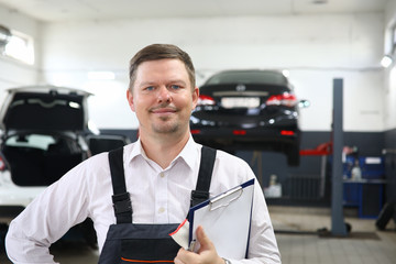 Portrait of laughing man holding important paper folder with information about different autos and standing in modern car maintenance garage. Machinery repairman concept